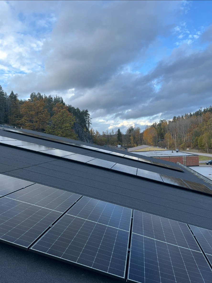 Solcellspaneler i långa rader på det mörka taket till ishallen i Valdemarsvik, med skogbevuxen häll i bakgrunden
